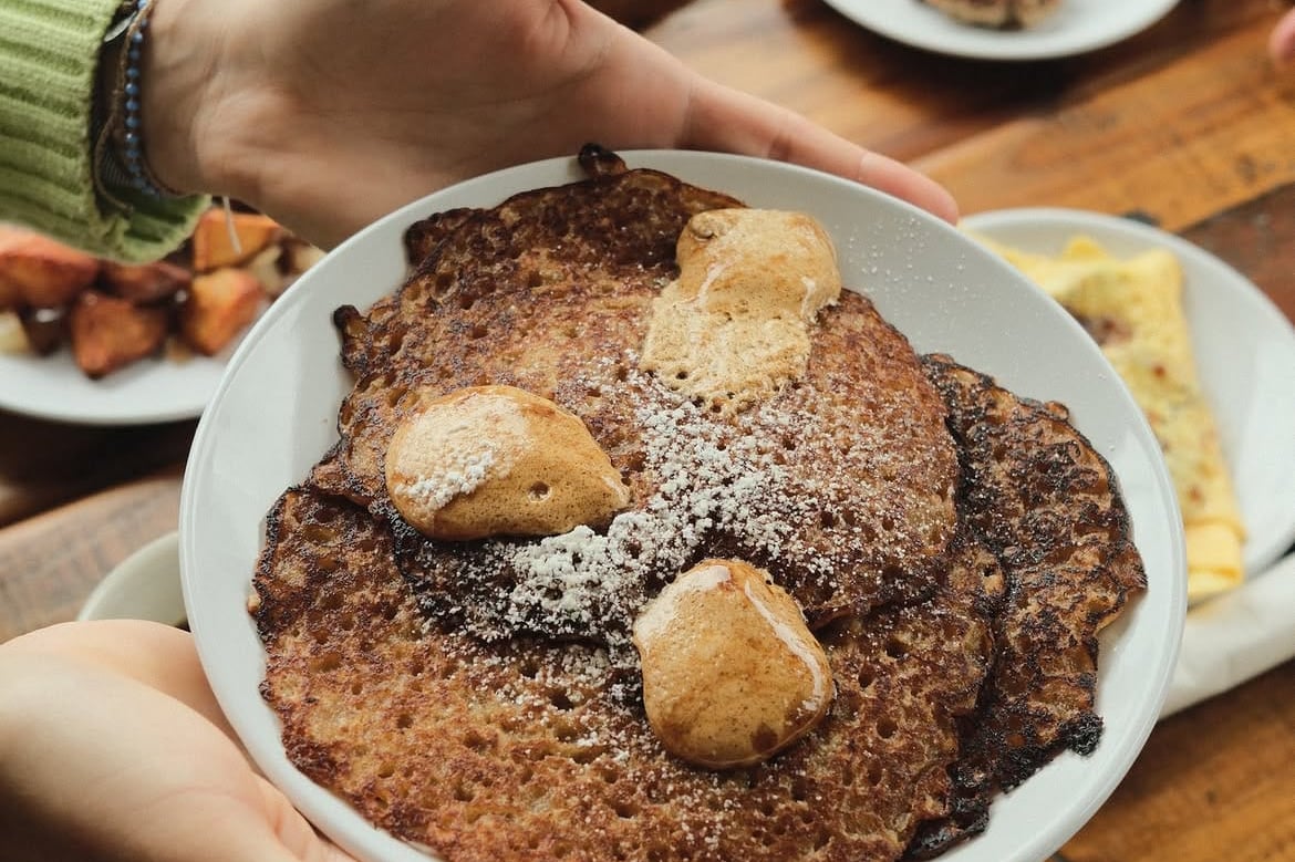 A plate of three oatmeal griddle cakes dusted with powdered sugar and topped with cinnamon molasses butter, held above a wooden table.