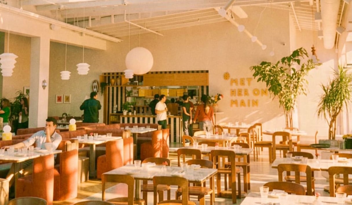 Sunlit dining room at Met Her at a Bar on Main Street in Santa Monica, with marble tables, orange banquettes and diners near the open kitchen.