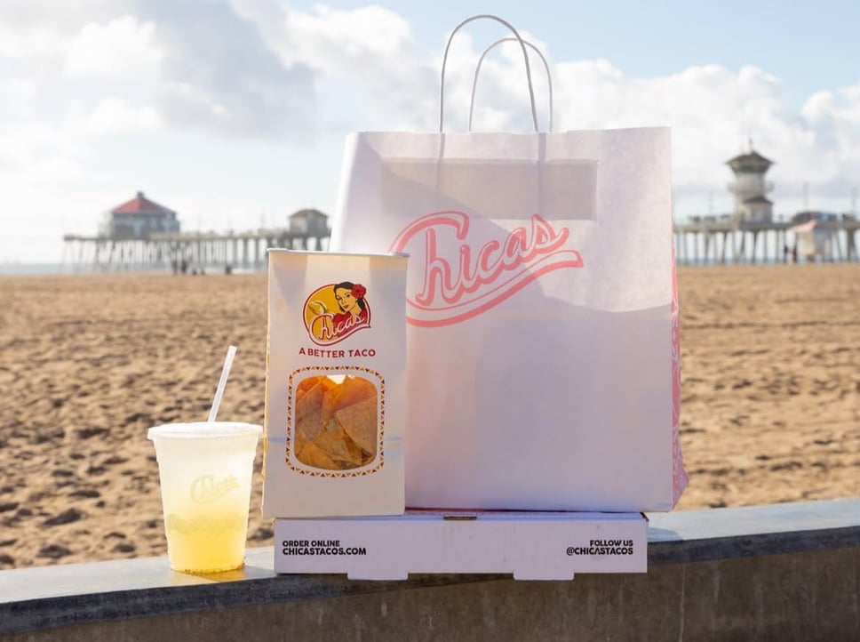 A Chicas Tacos paper bag, a carton of tortilla chips and an agua fresca sitting on a low wall above the sand, with a pier in the background