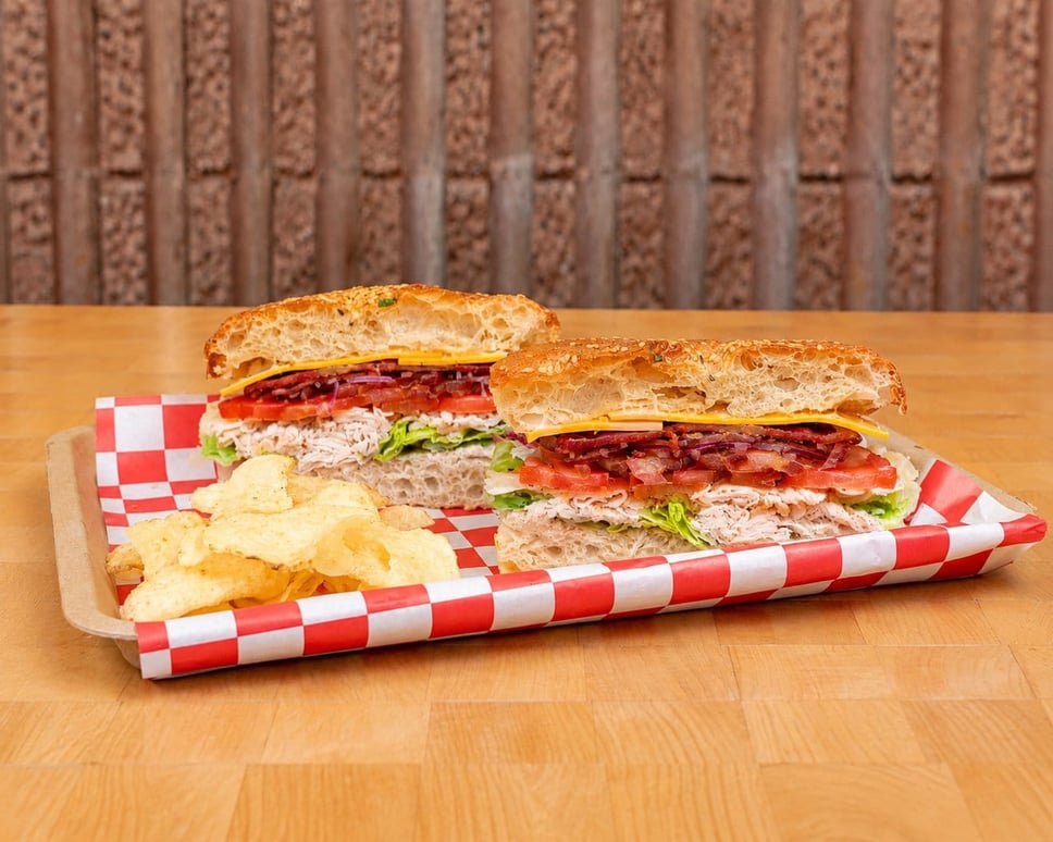 Two halves of the Nostalgia club sandwich with turkey, candied bacon, American cheese, romaine and tomato on sesame focaccia, served with potato chips on a red checkered tray