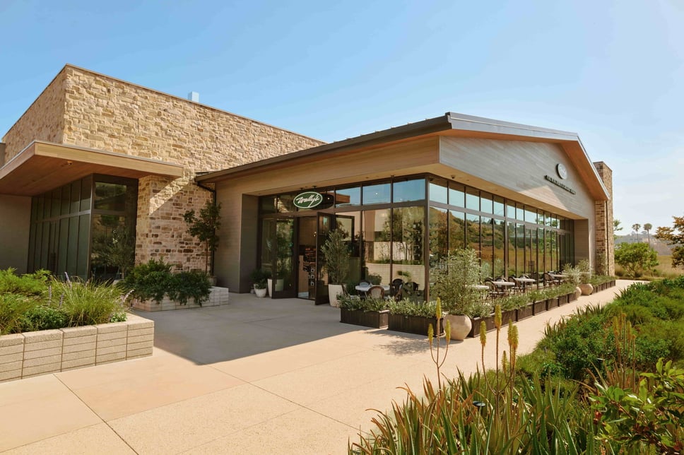The exterior of Wally's Malibu at Cross Creek Ranch, a stone and timber building with floor-to-ceiling windows, patio seating and drought-tolerant landscaping under a clear sky