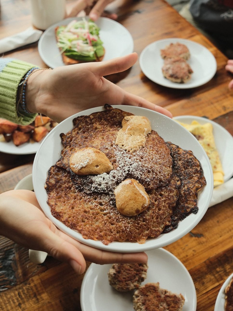 Two hands presenting a plate of oatmeal griddle cakes with lacy crisp edges, three quenelles of whipped butter and a dusting of powdered sugar