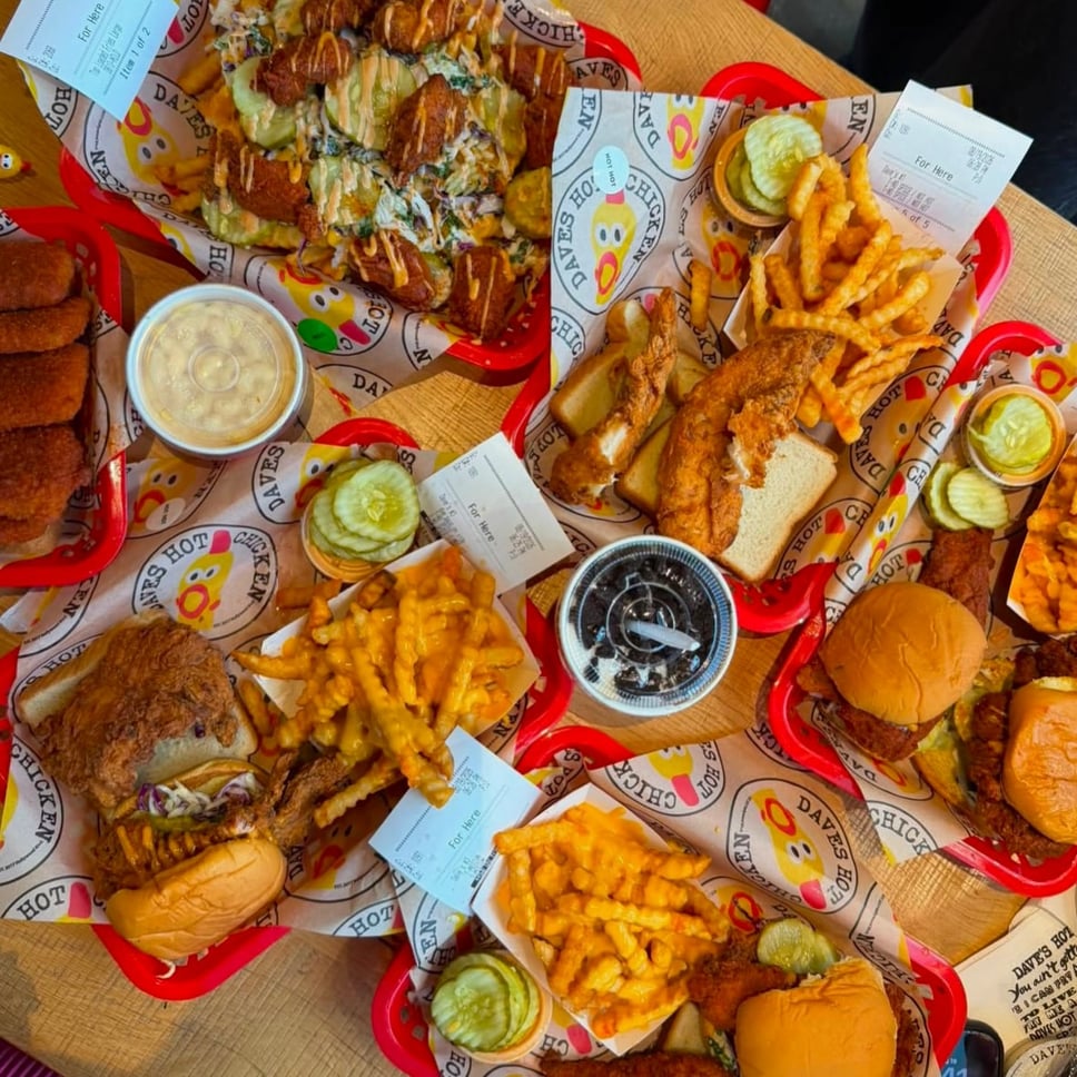 A table covered in Dave's Hot Chicken red baskets with branded liners, holding tenders, sliders, cheese fries, mac and cheese, pickles and a shake