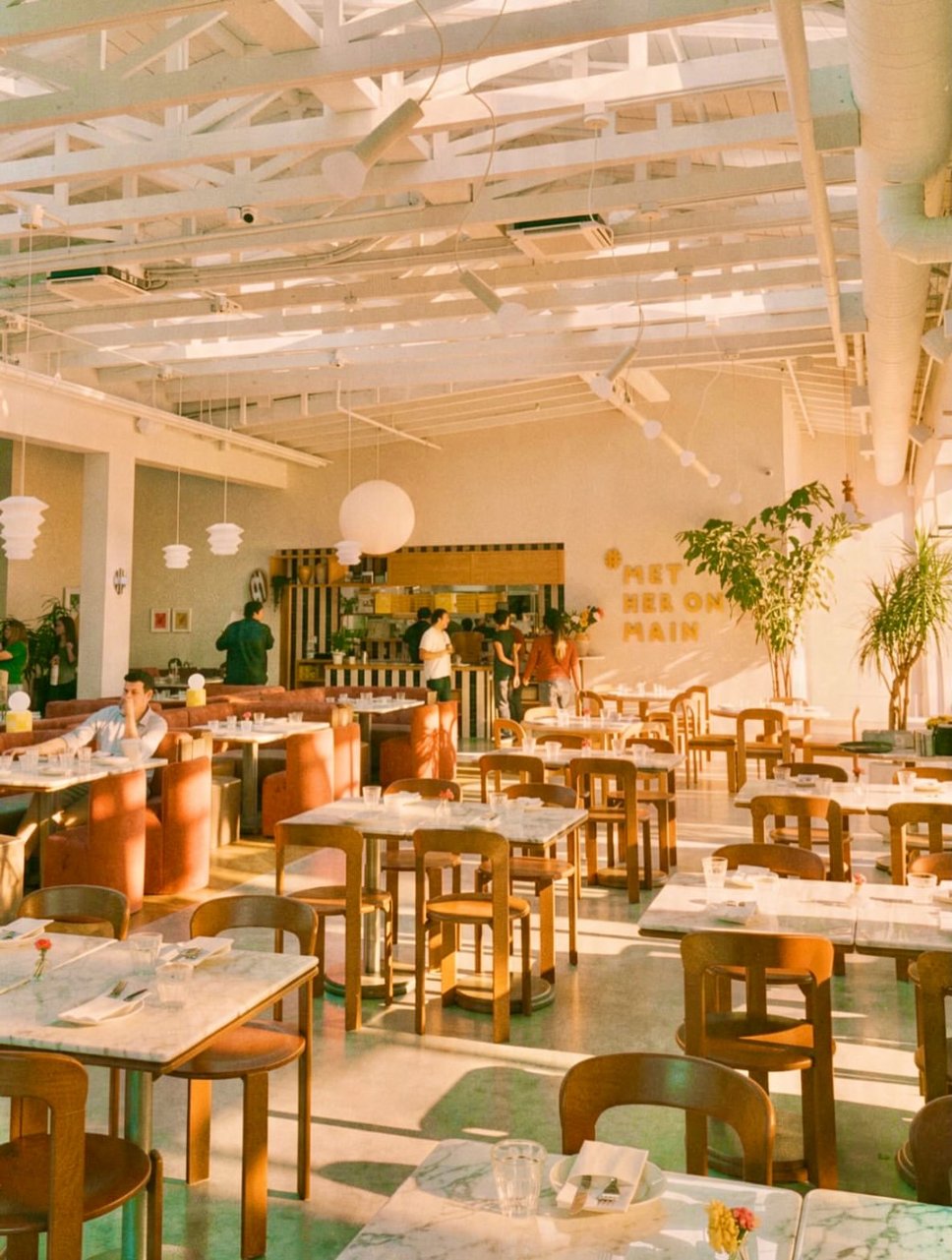 The sunlit Met Her at a Bar dining room on Main Street, with marble tables, rust velvet booths, paper lanterns and gold letters on the wall reading Met Her on Main