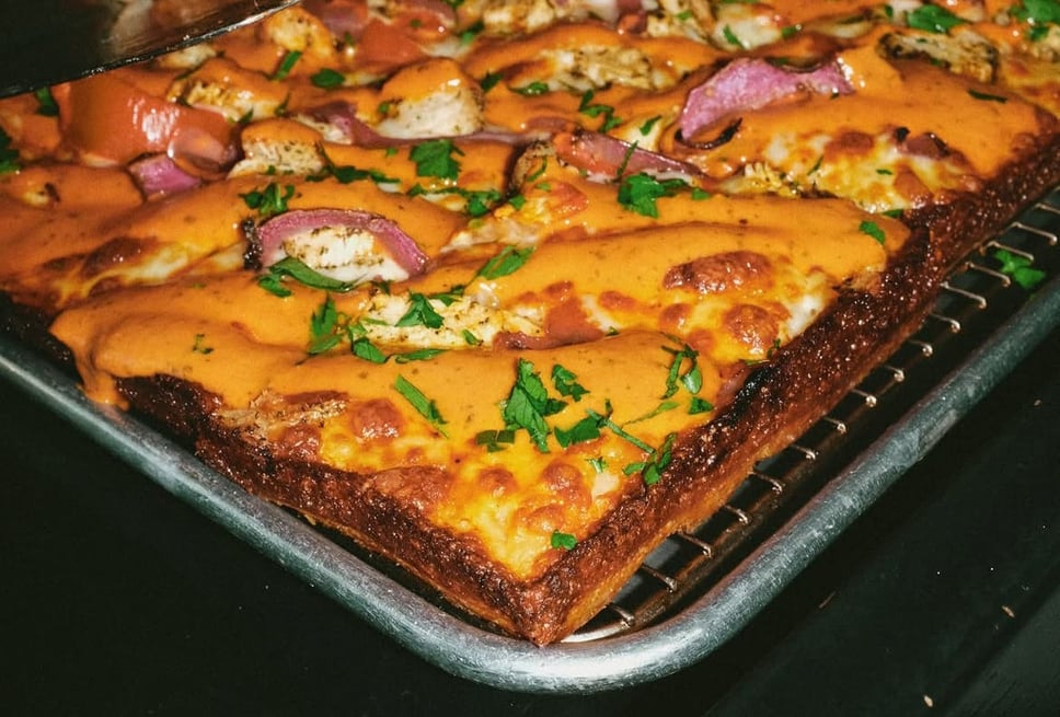 A rectangular Prime Pizza pan pie cooling on a wire rack, covered edge to edge in a creamy orange sauce with chicken, red onion, bell pepper and chopped parsley