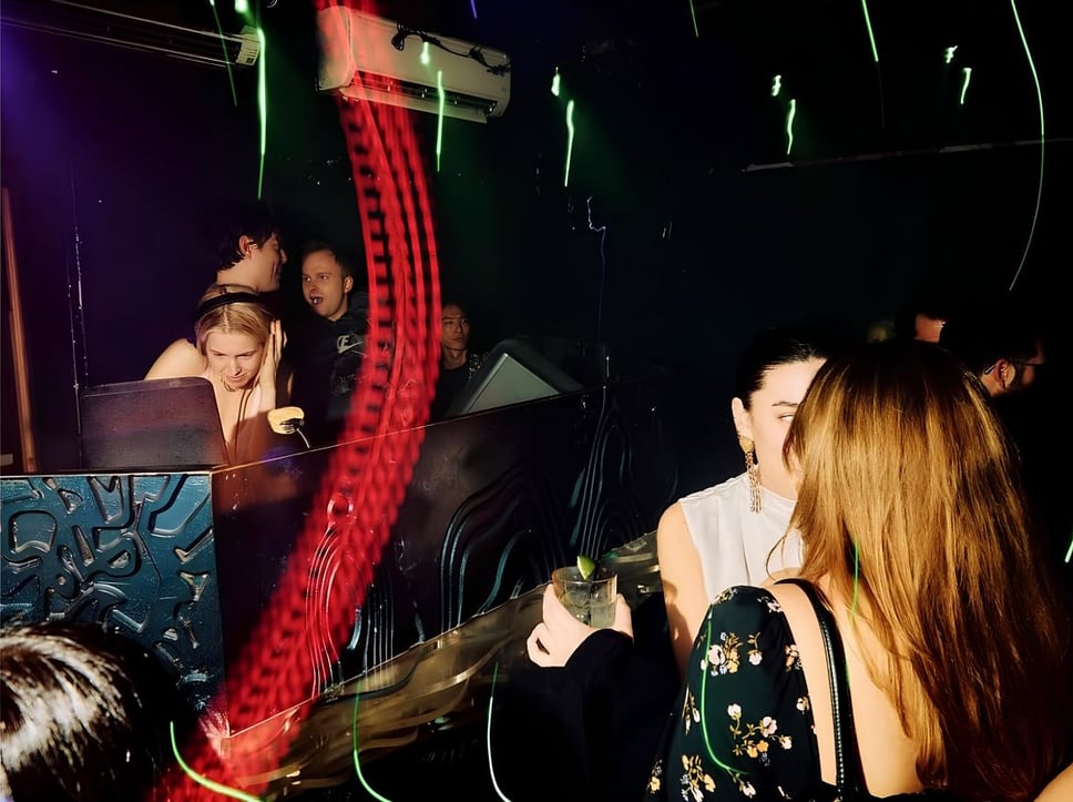 Two female patrons talking to one another in a candid-like retro shot with a DJ in the DJ booth spinning tracks, three men in the back behind the DJ talking to one another
