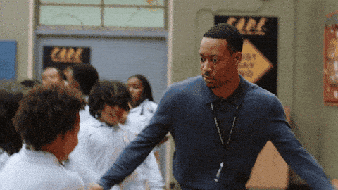 A teacher in a blue sweater and lanyard dances in a line with students at a school dance.
