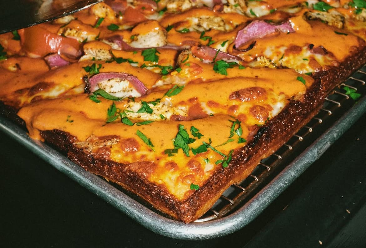Flash-lit close-up of a square butter chicken pan pizza with melted cheese, pickled red onions and chopped herbs on a metal tray.