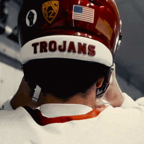 A USC Trojans player pulls on a cardinal helmet with TROJANS across the back.