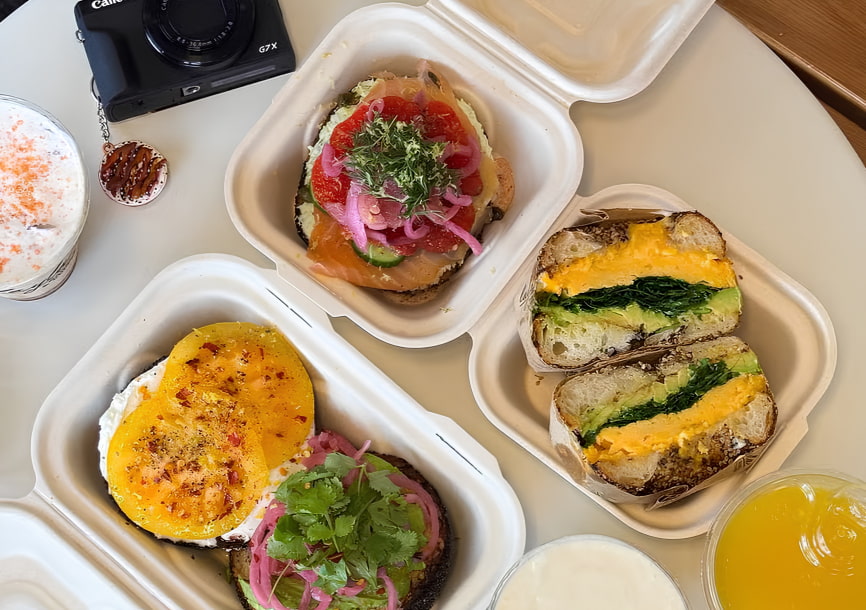 Assorted open-faced and halved bagel sandwiches in takeout boxes, with drinks on a table.