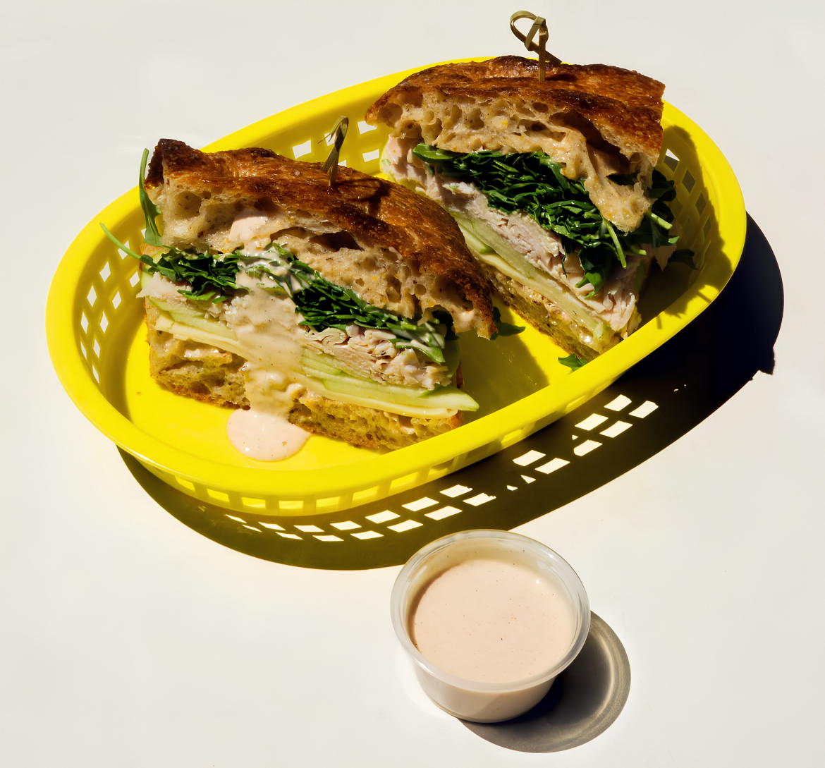 Two sandwich halves with sliced meat, cheese, and leafy greens in a yellow basket, with sauce on the side.