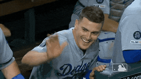 A smiling Dodgers player waves from the dugout, then turns toward his teammates.
