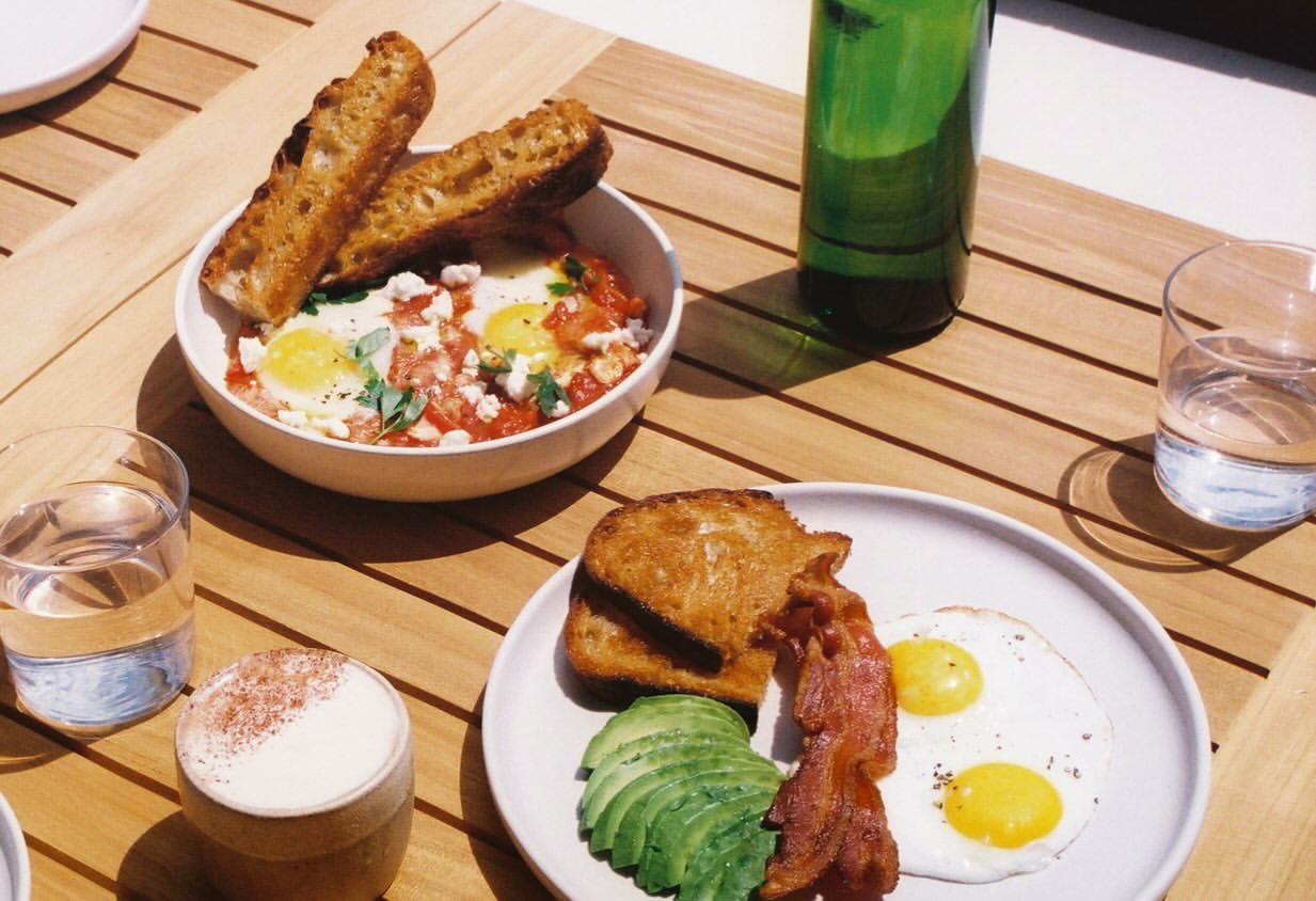Breakfast plates with eggs, bacon, avocado and toast, plus coffee, on a wooden table at Bower Surf Club.