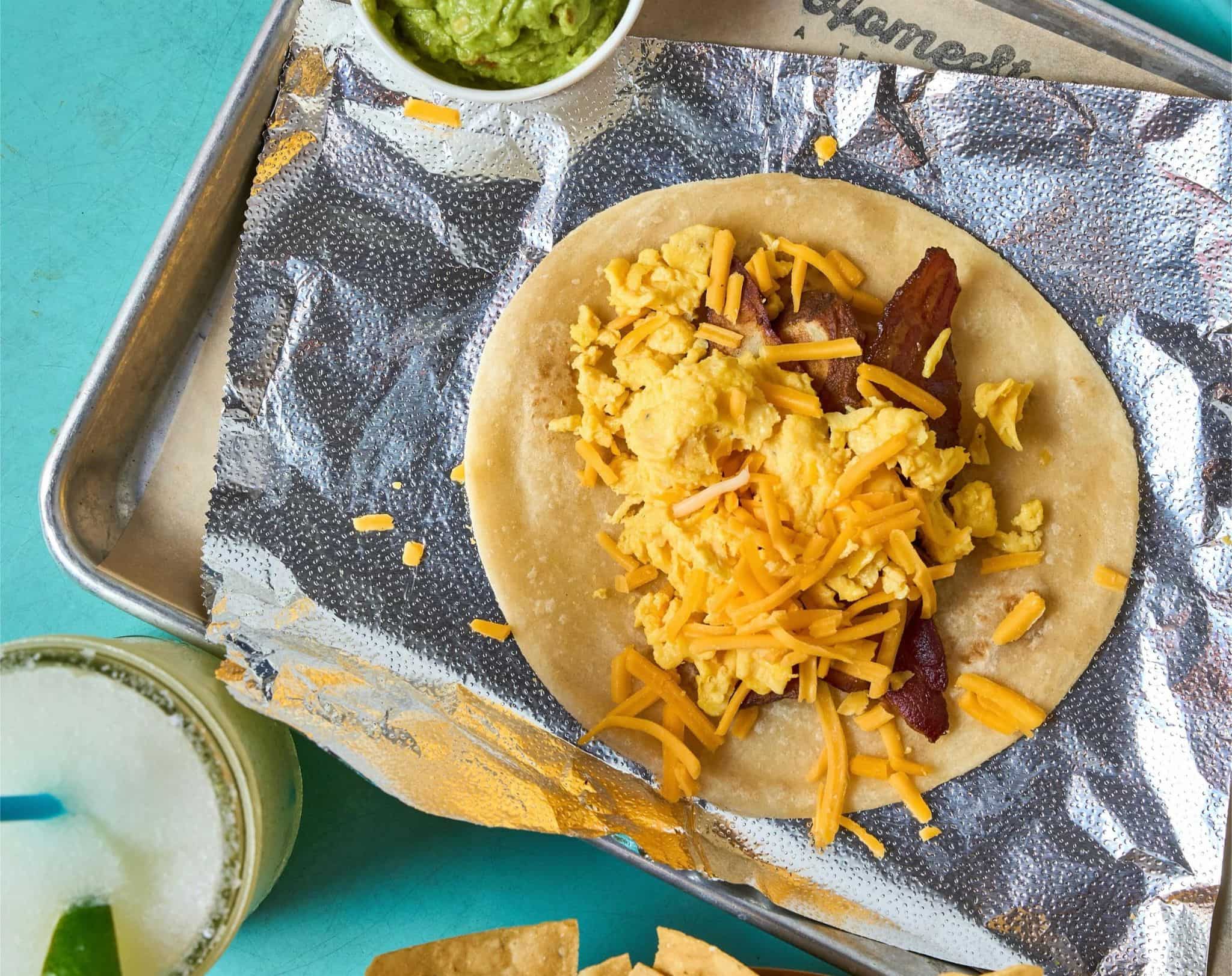 HomeState breakfast taco topped with eggs, bacon, and cheddar, beside guacamole and lime.
