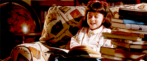 Matilda laughing with delight while reading, surrounded by stacks of books.